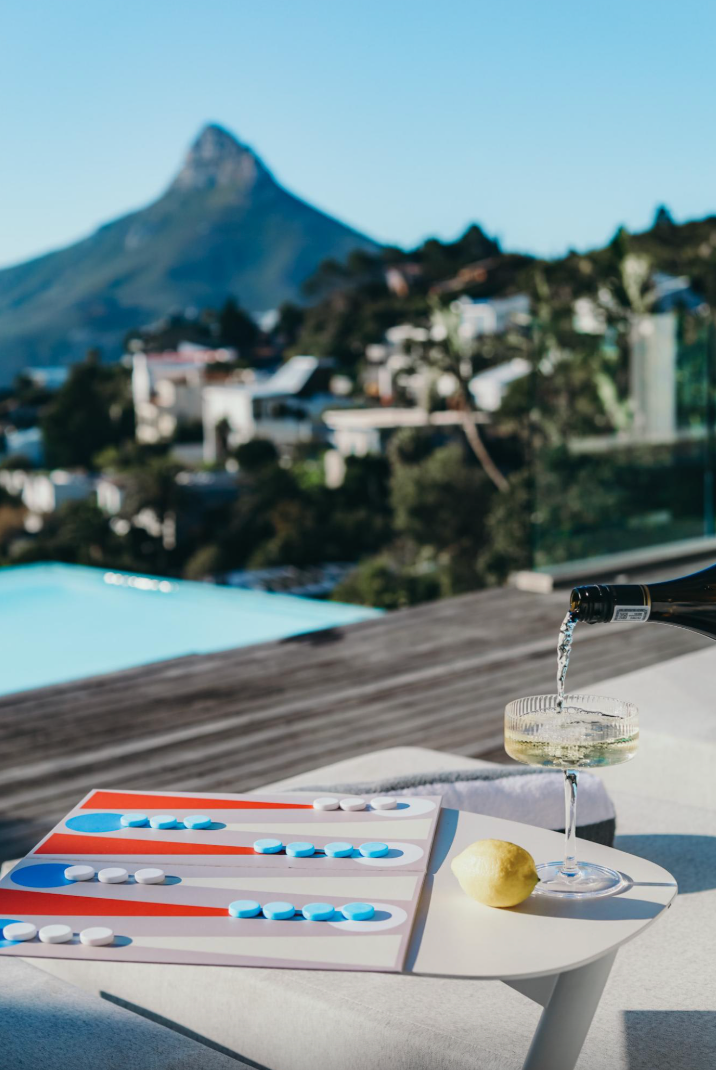 A hand pours sparkling wine into a glass on a table with a board game and lemon, overlooking a mountain.