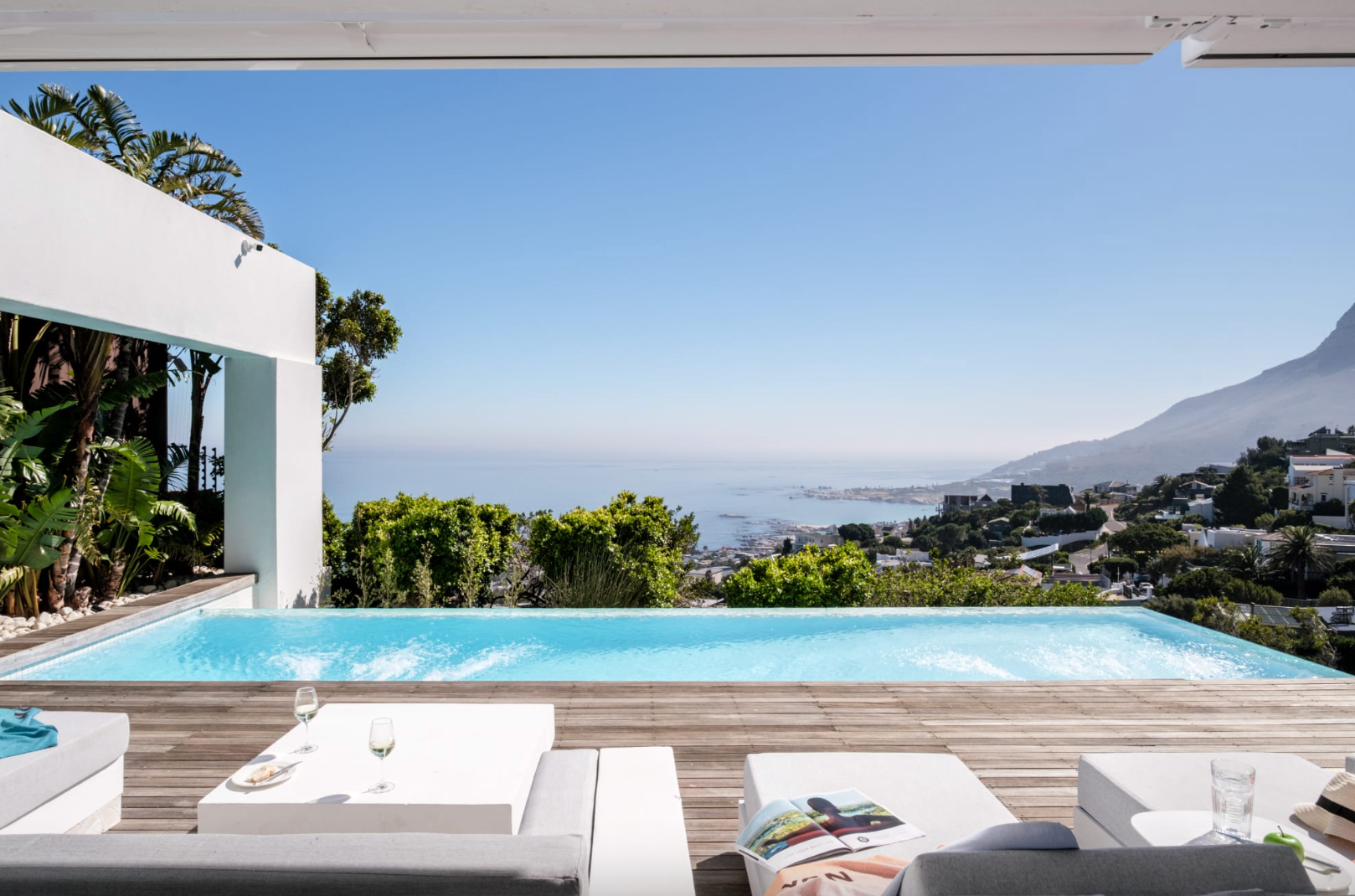 Infinity pool overlooking a city and ocean, with white lounge furniture and a blue sky.