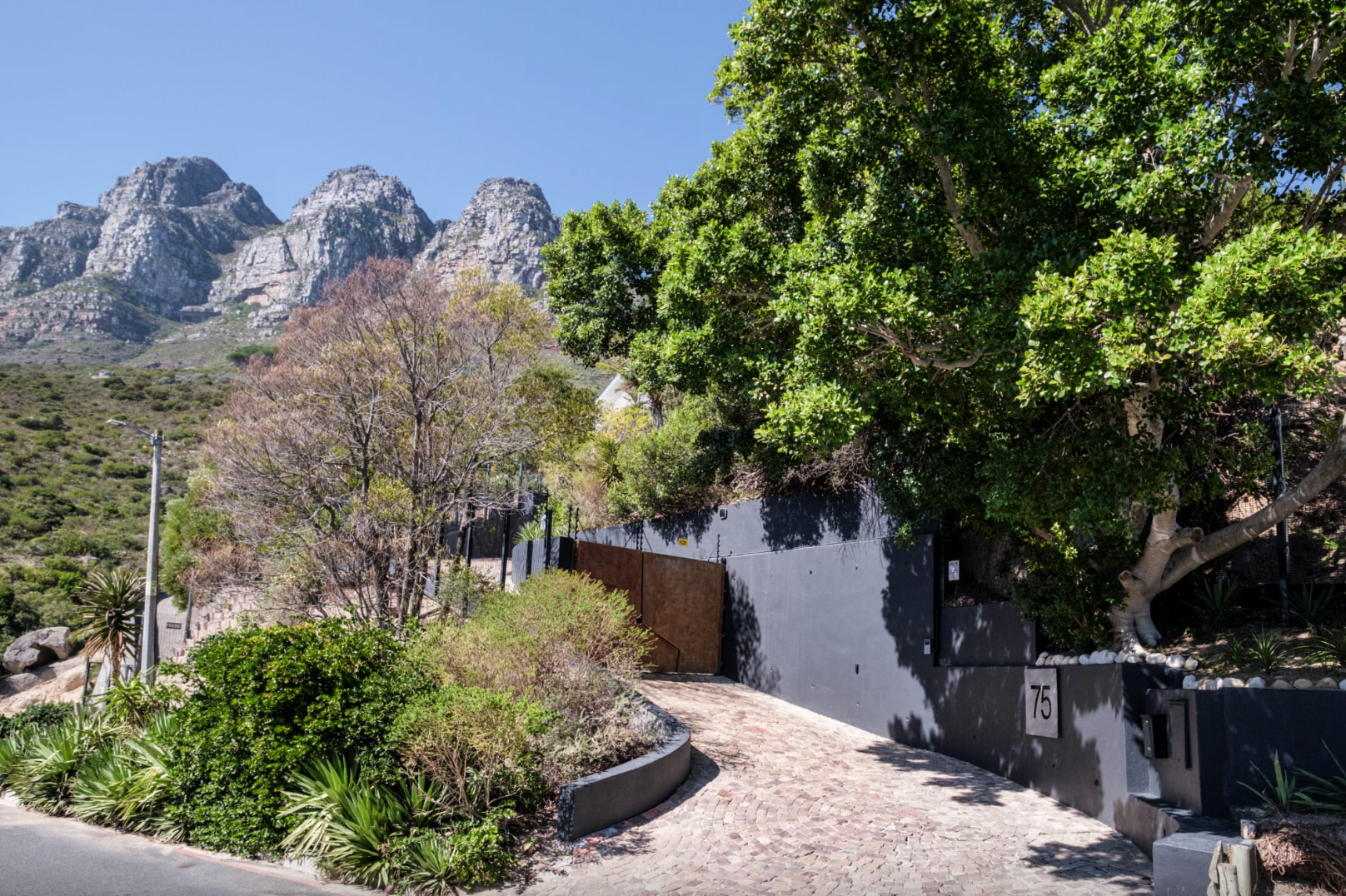 Driveway with gray walls, wooden gate, and mountains in background. Lush trees and foliage frame the entrance.