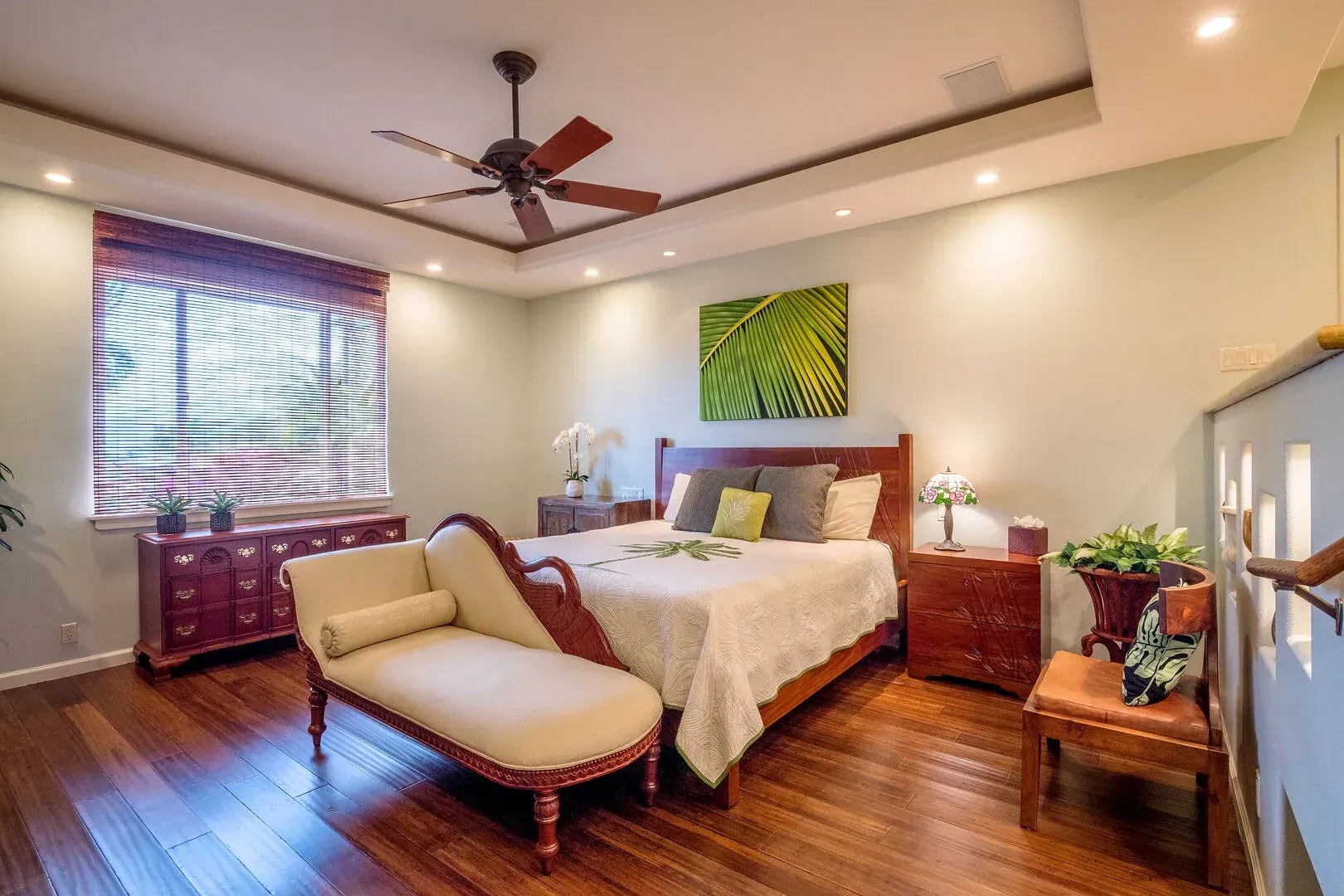 A bedroom with hardwood floors , a bed , a chaise lounge and a ceiling fan.