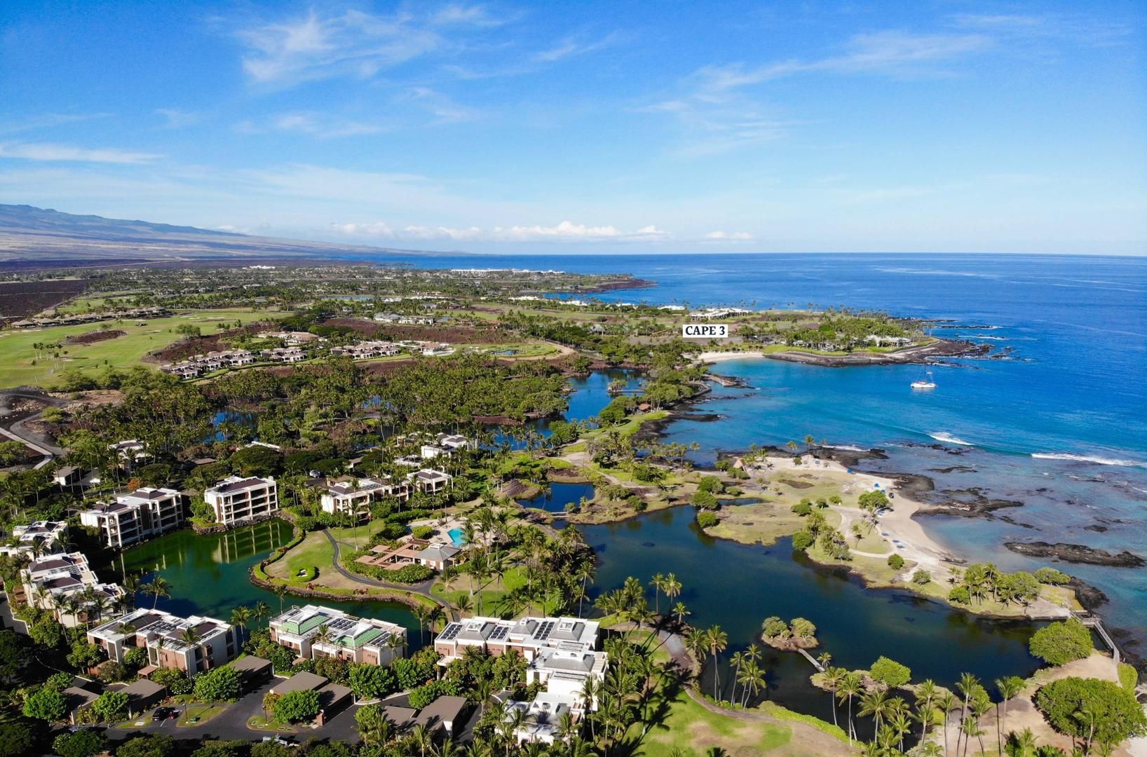 Aerial view of a coastal resort with buildings, lagoons, greenery, and the blue ocean under a bright sky.