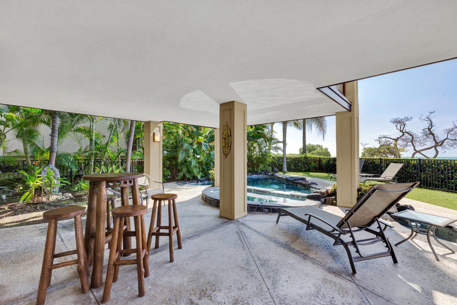 Patio with wooden stools and high table. Lounge chair by a pool, lush greenery, and blue sky.