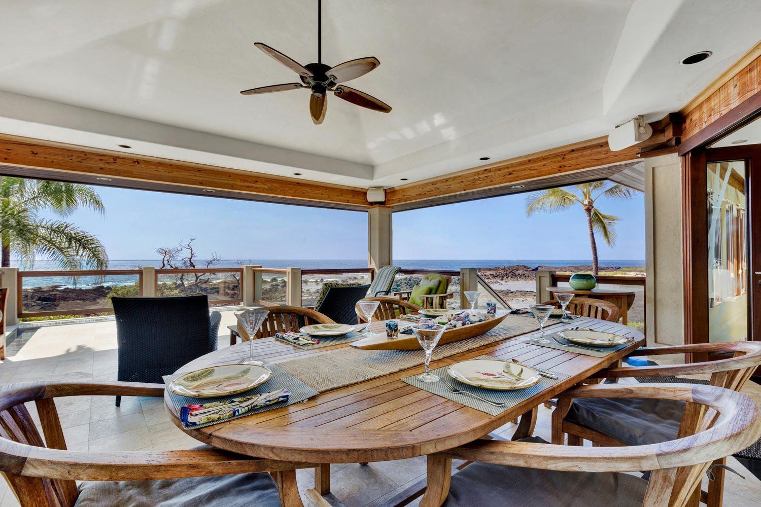 Outdoor dining table with ocean view; wooden chairs, table setting, ceiling fan.