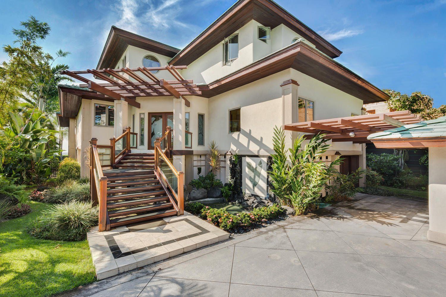 Two-story beige house with wooden accents, a walkway, and greenery, under a bright blue sky.