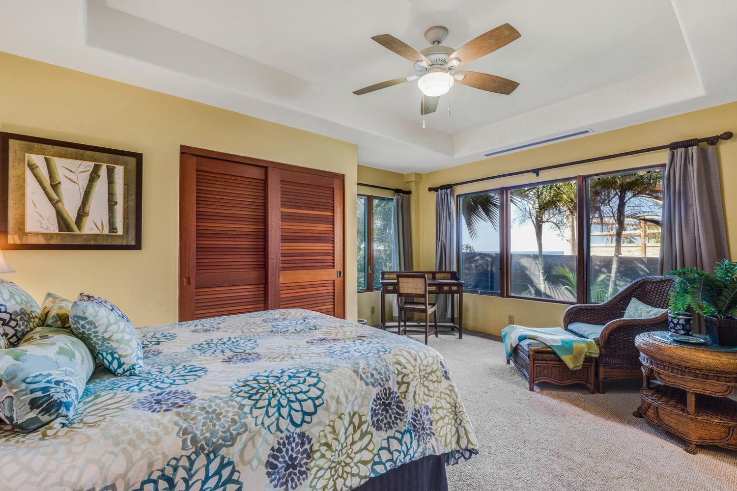 Bedroom with bed, wooden doors, windows, desk, wicker chair, and beige carpet.