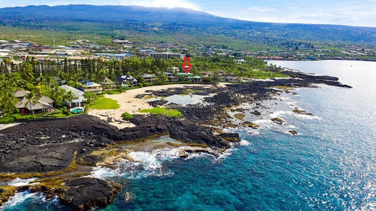 Coastal view of a beach with black lava rocks, turquoise water, and a red tower in the background.