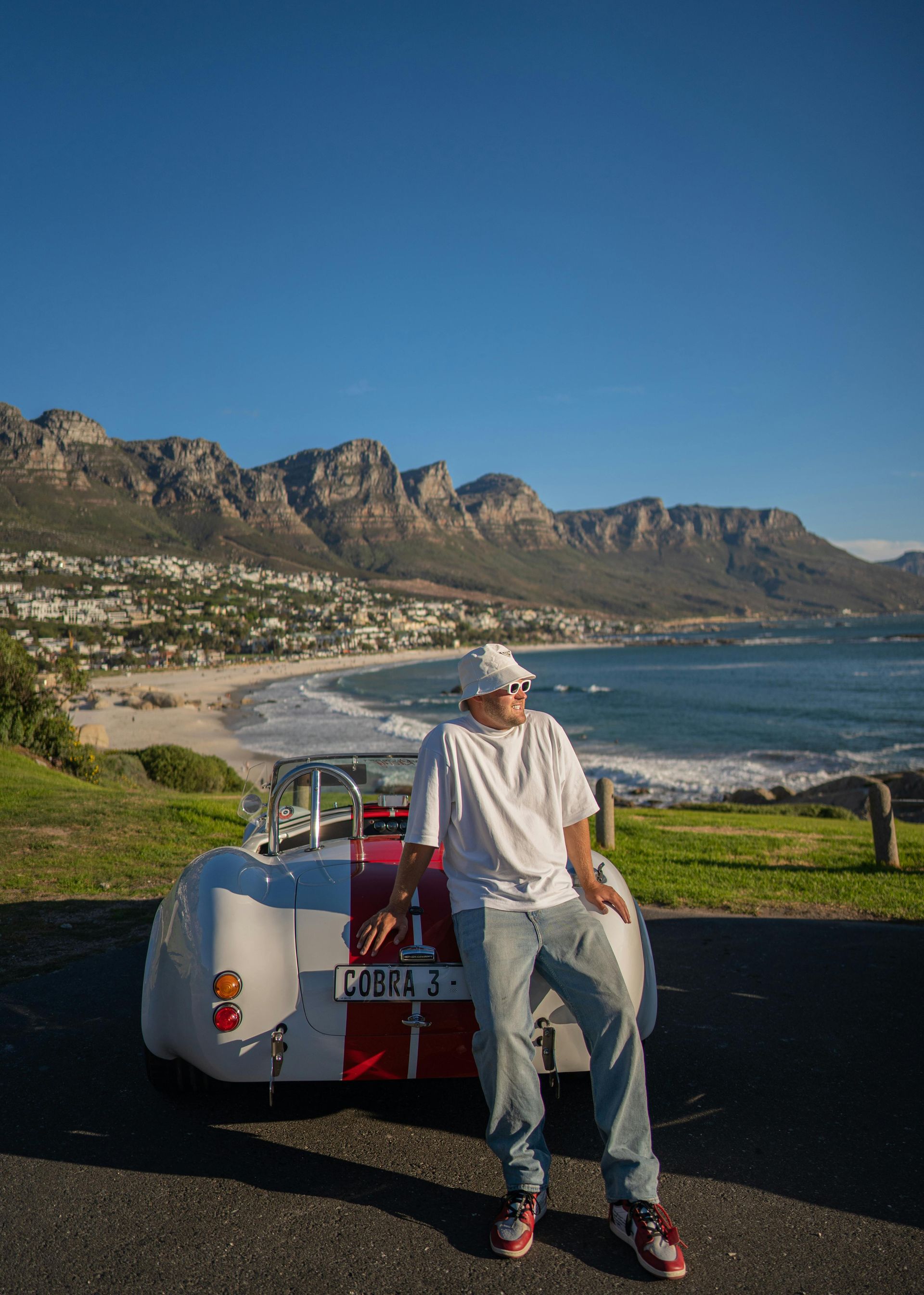 Person leaning on a white convertible car overlooking a beach and mountain range in Cape Town.