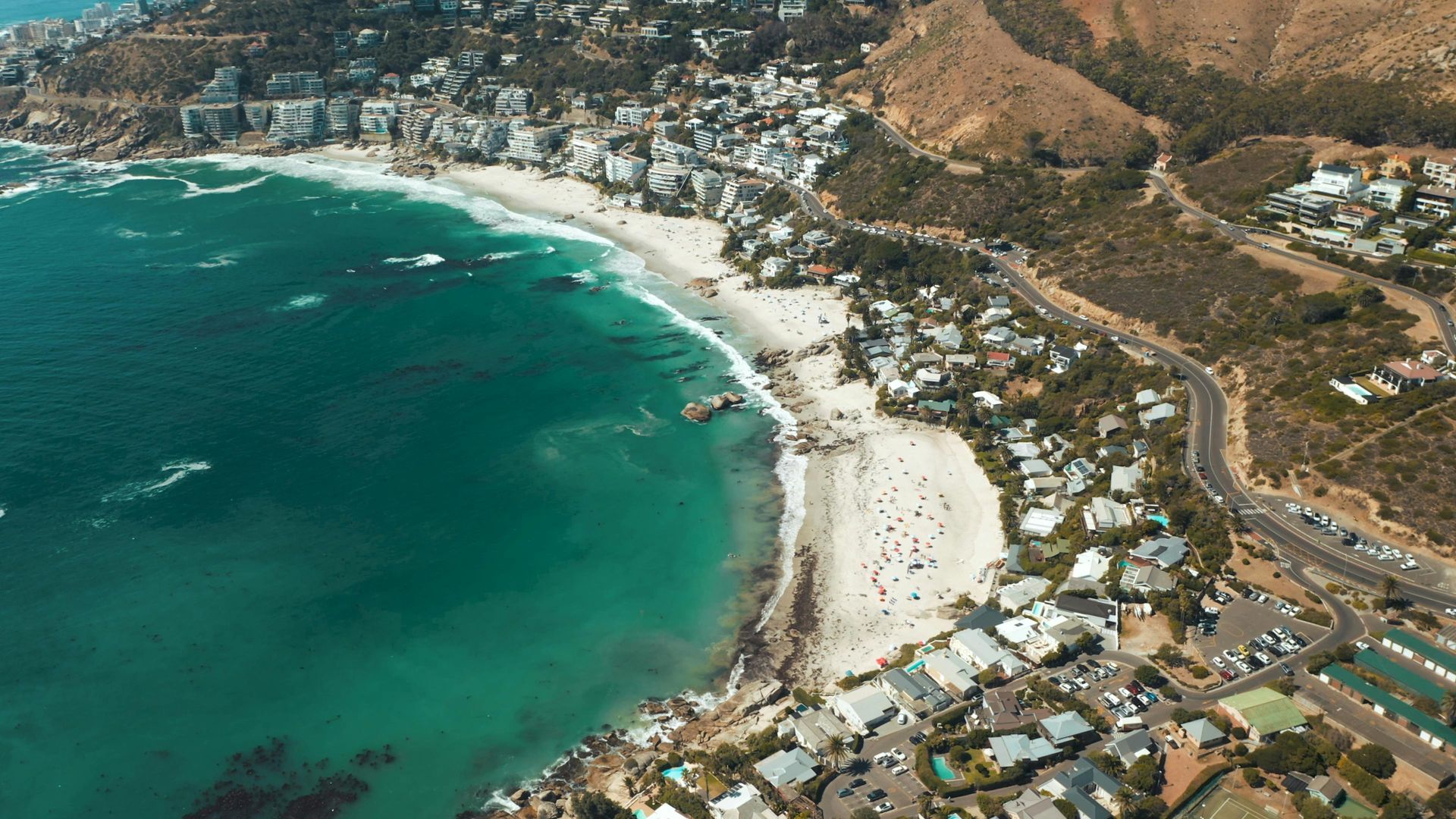 Aerial view of a beach with turquoise water, a sandy shore, and buildings along the coast under a sunny sky.
