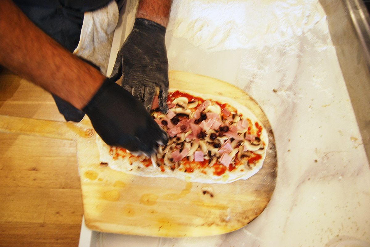 A man wearing black gloves is putting toppings on a pizza