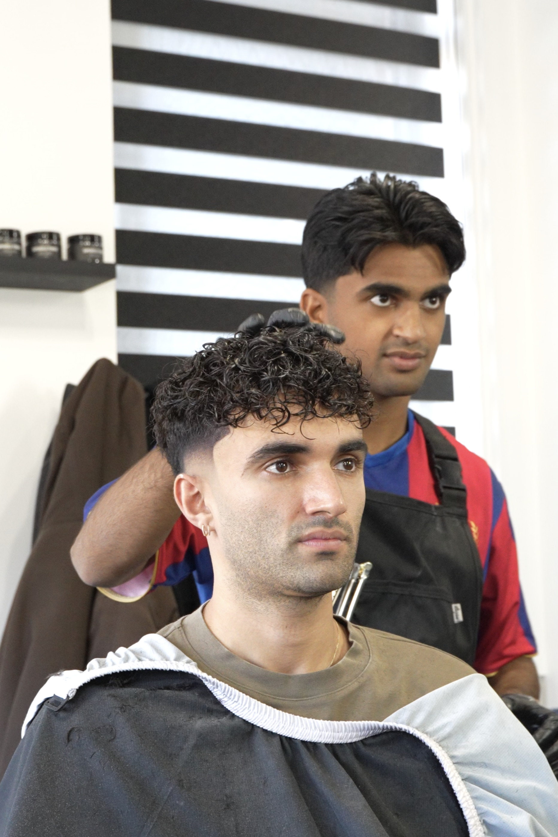 A barber standing behind a man, finishing a haircut with a fade and curly top in a barbershop.