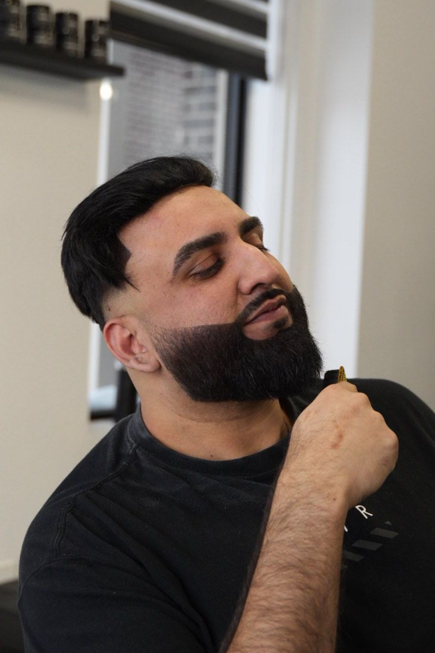 A man with a neatly groomed, dark beard and a short fade haircut holds a hair trimmer while looking away.