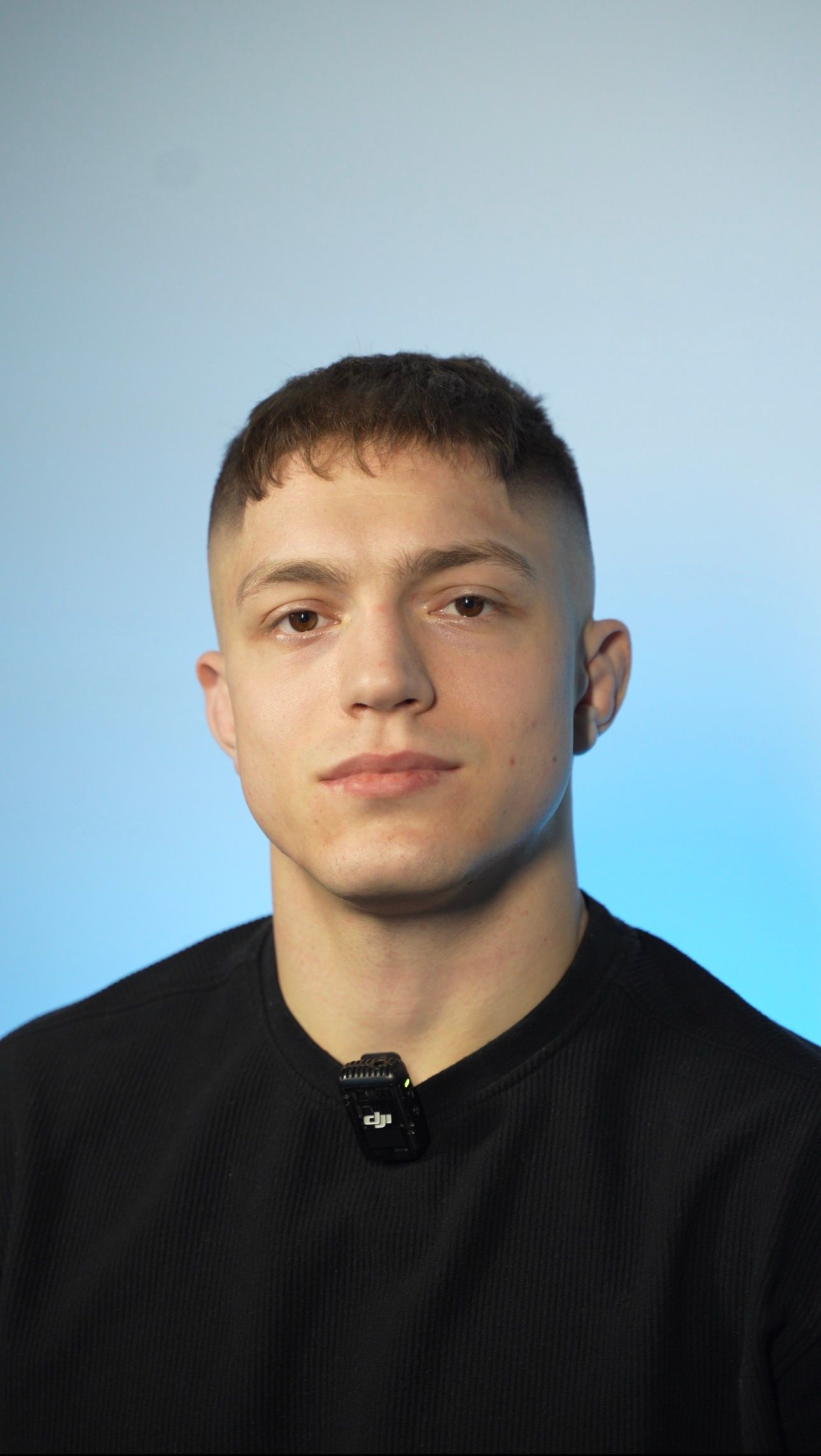 A young person with short, dark hair wearing a black shirt, centered against a light blue background.