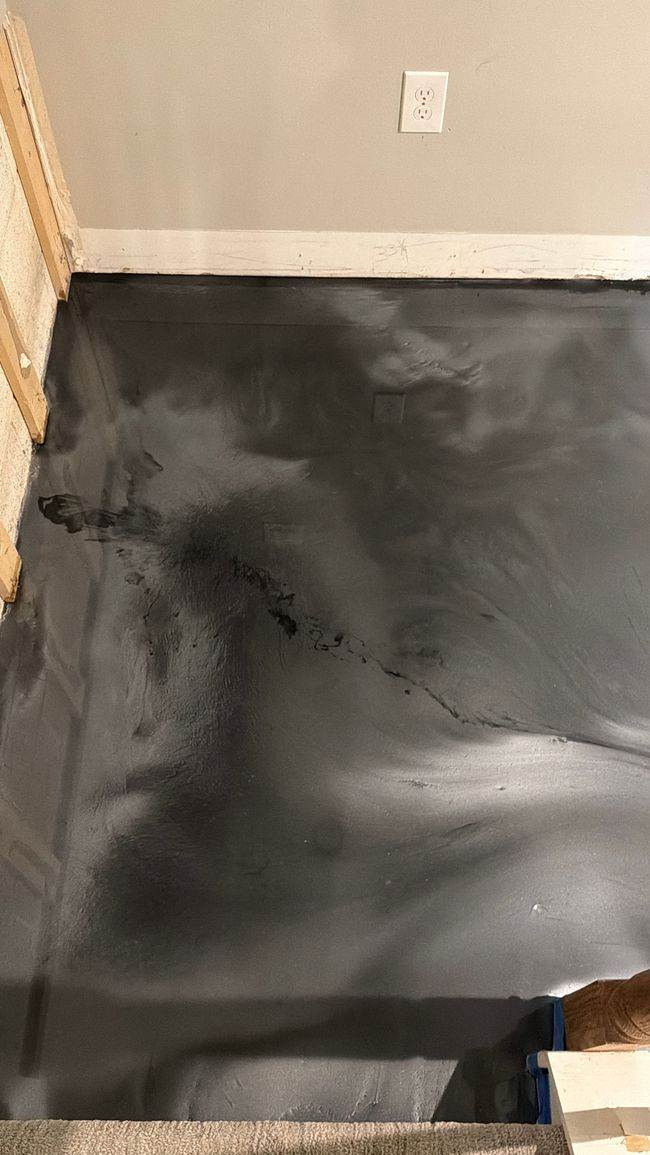Black floor surface with wet patches near a beige wall and electrical outlet.