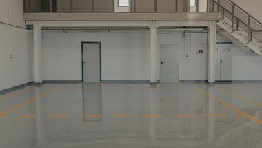 Empty indoor warehouse with glossy floor, white walls, stairs, and a small door under a mezzanine.