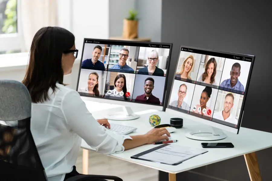 Woman in glasses at a desk on a video conference call with many other participants on two monitors.