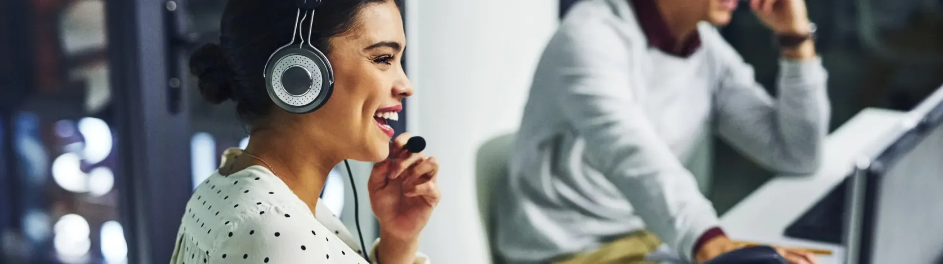 Woman wearing headset, smiling while speaking. Man in background, working at a desk.