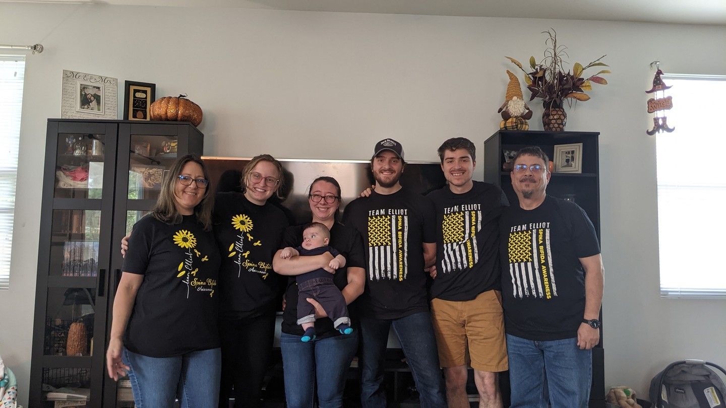 A family of seven stands in front of a wall, some wearing matching shirts.