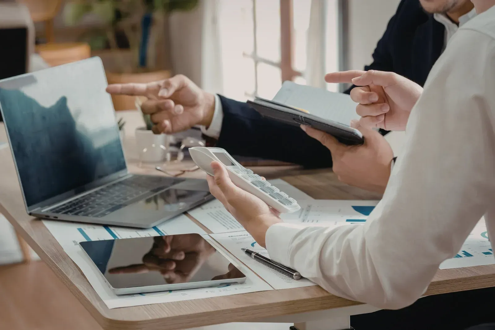 People in business attire working at a table, using a laptop, tablet, calculator, and documents with charts.