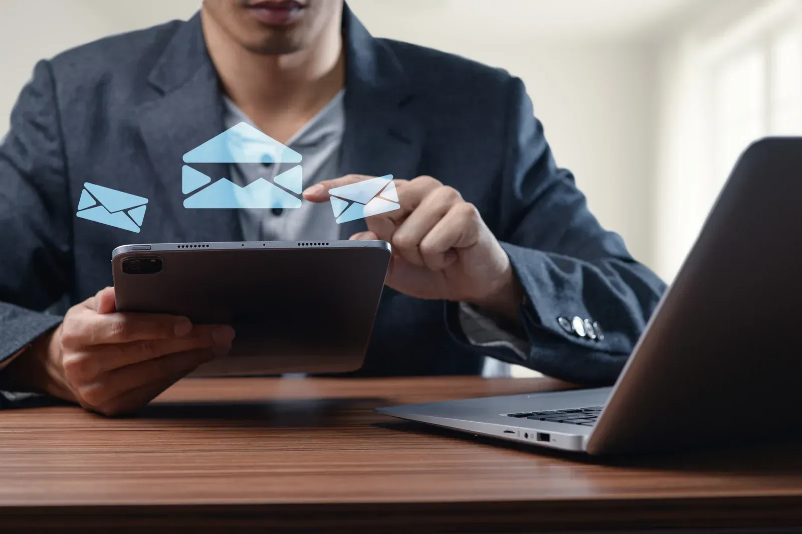 Man in blazer using tablet, interacting with digital email icons, near laptop on desk.