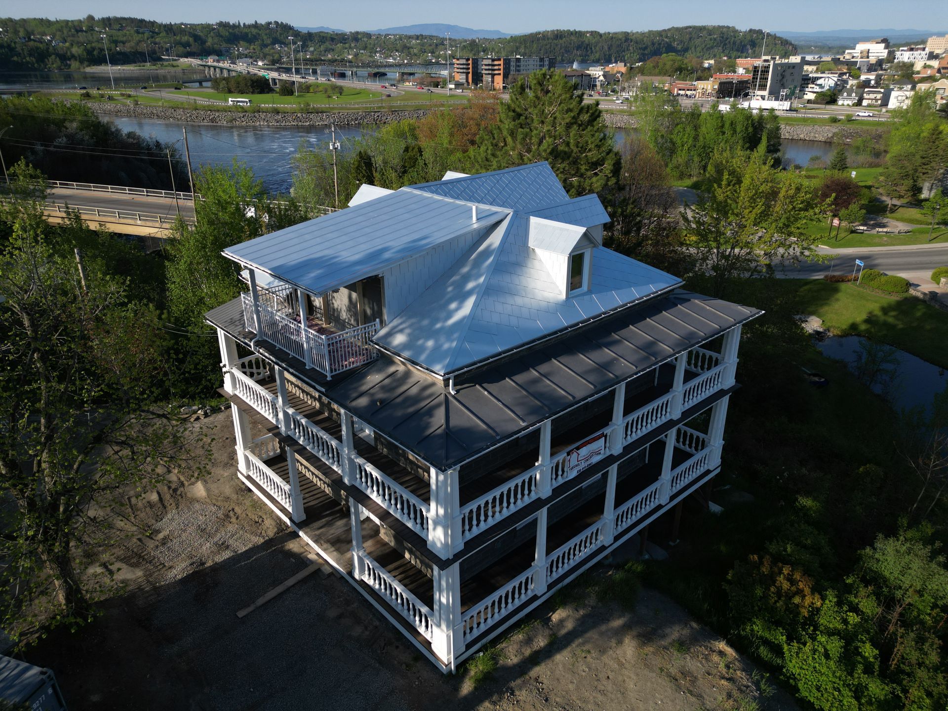 Maison blanche de trois étages avec vérandas enveloppantes et toit argenté donnant sur l'eau et une ville.