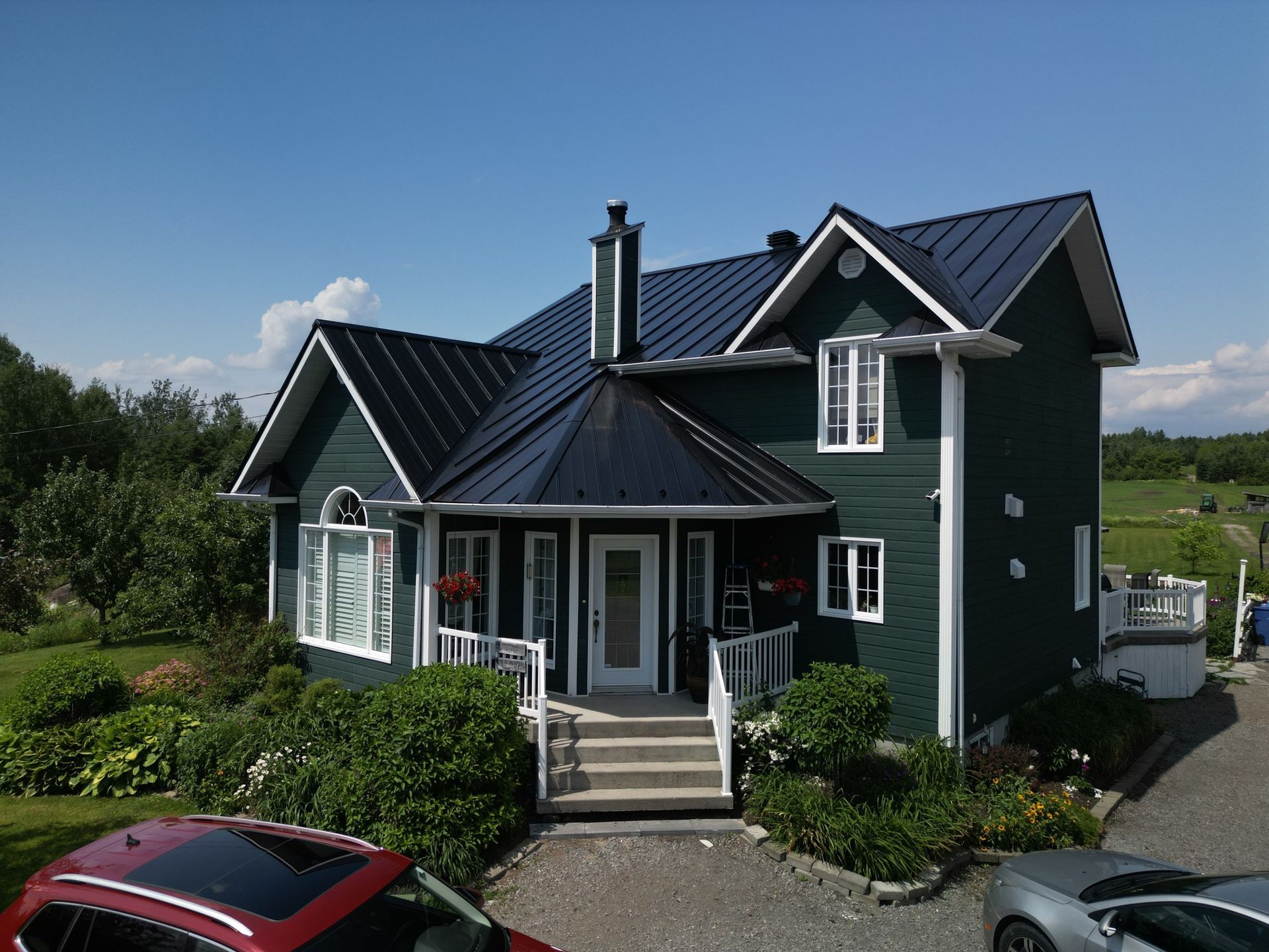 Maison verte au toit noir et aux boiseries blanches, porche d'entrée et voitures garées dans l'allée par une journée ensoleillée.