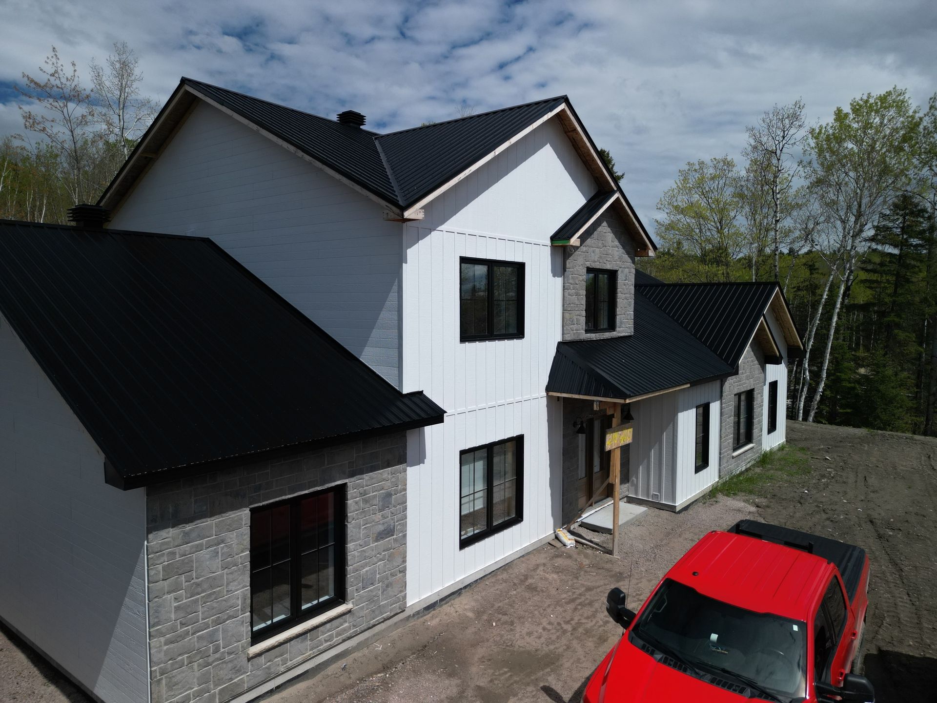 Maison moderne à deux étages avec toit noir, bardage blanc et éléments en pierre grise. Un camion rouge est garé dans l'allée.