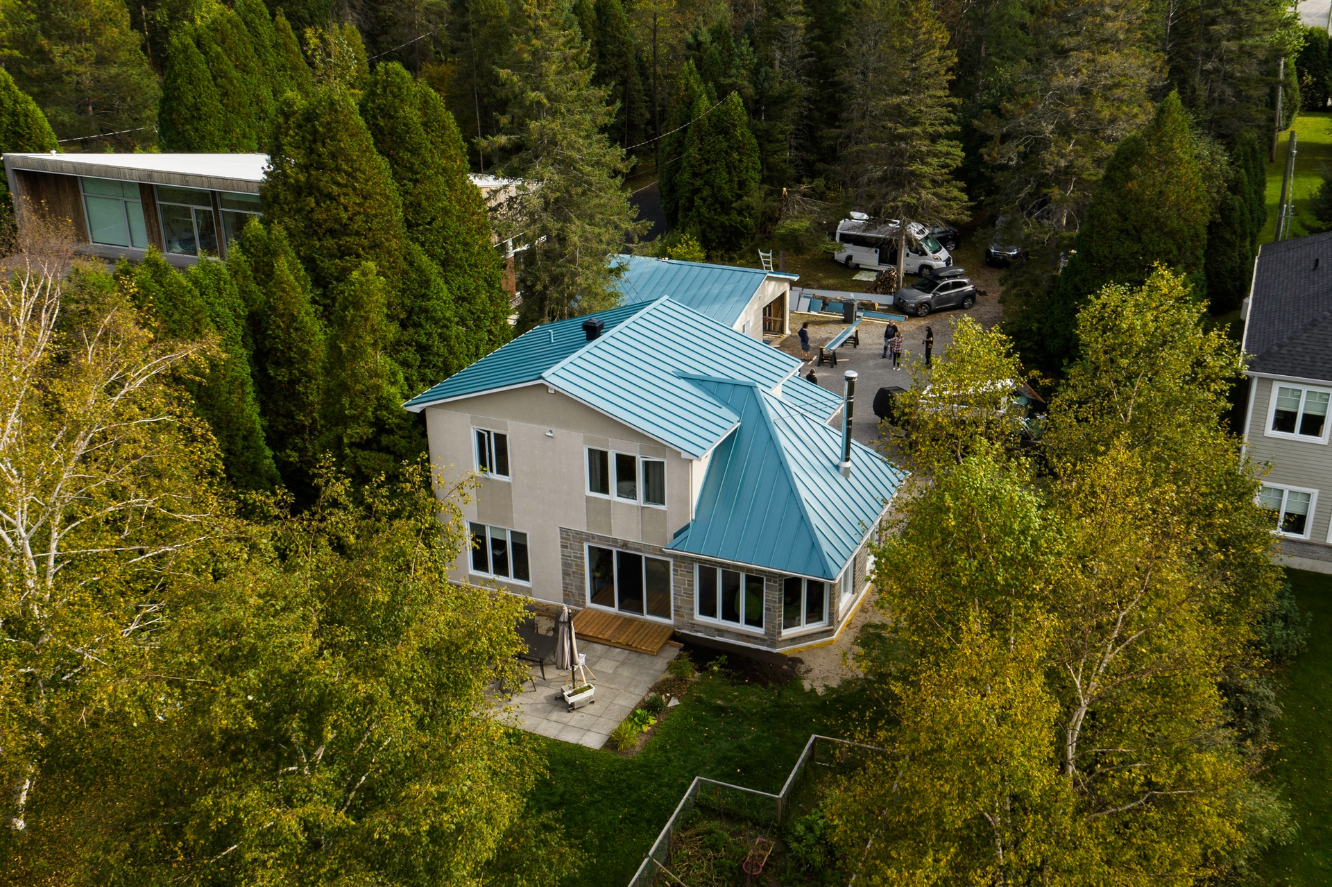 Vue aérienne d'une maison à deux étages au toit bleu, entourée d'arbres et d'autres maisons.