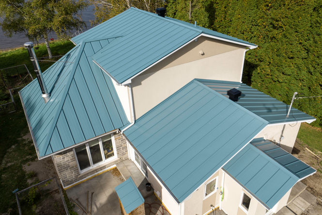 Toiture métallique bleue sur une maison à deux étages avec bardage beige, cheminée et arbres verts environnants.