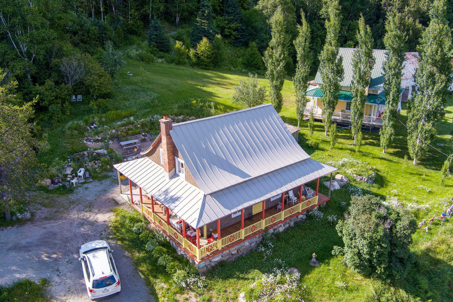 Cabane avec toit en métal et véranda enveloppante, voiture garée, autres cabanes au loin, entourée d'arbres et de verdure.