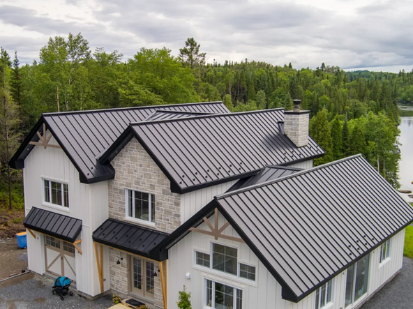 Maison blanche à deux étages avec toit en métal gris foncé, façade en pierre et éléments en bois, située dans une zone boisée près de l'eau.