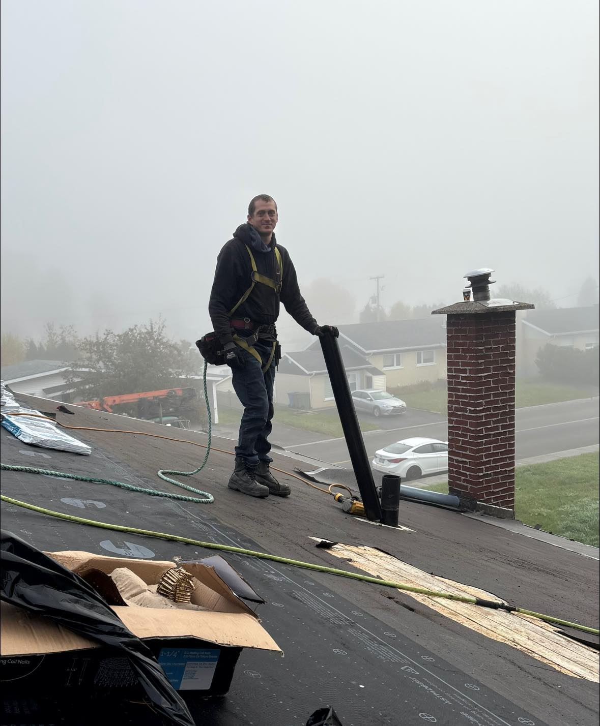 Couvreur sur un toit enveloppé de brouillard, tenant un tuyau près d'une cheminée. Harnais de sécurité sur la tête, il sourit.