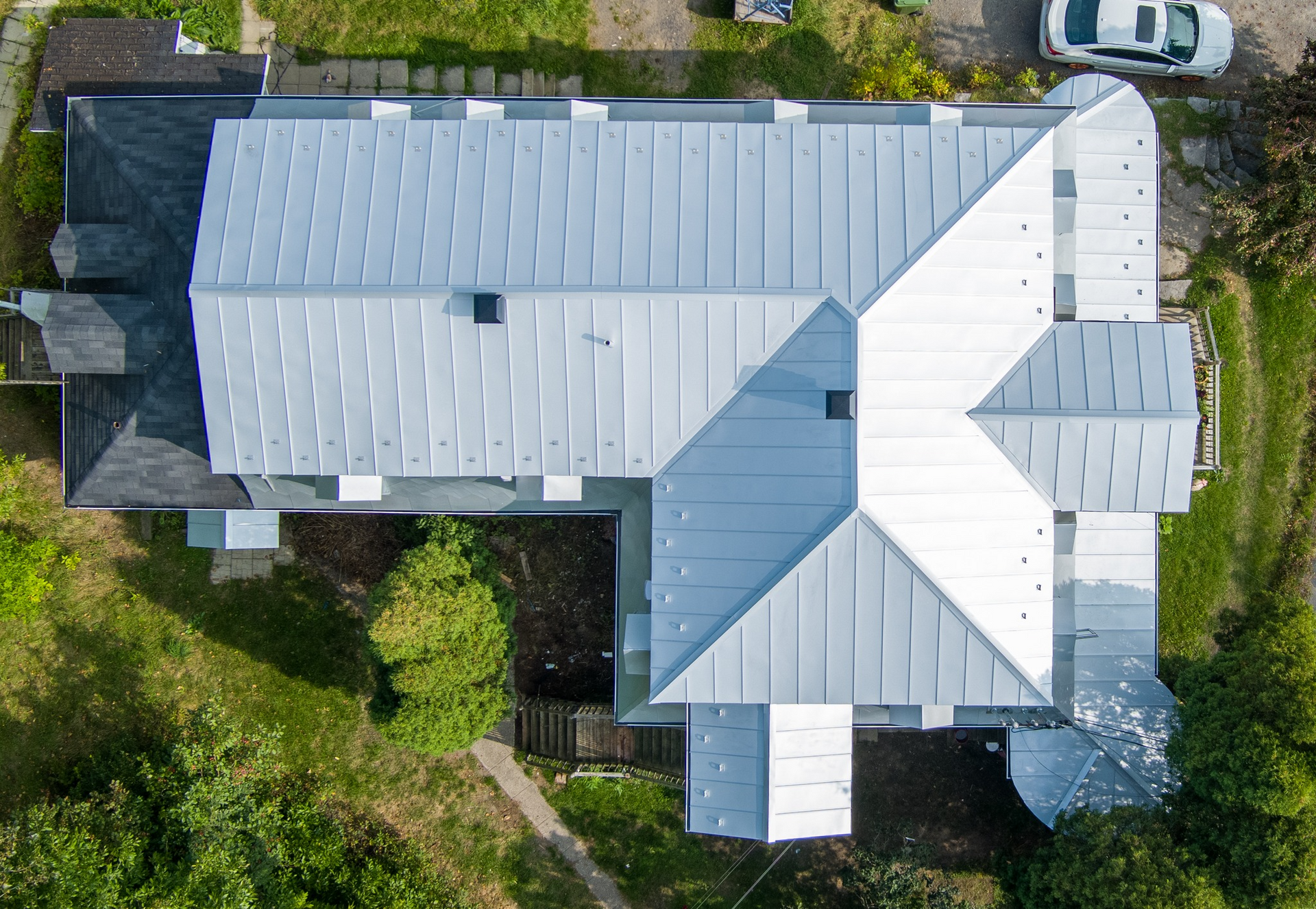Vue aérienne d'une maison au toit en métal gris, entourée d'herbe verte et d'une petite voiture blanche.