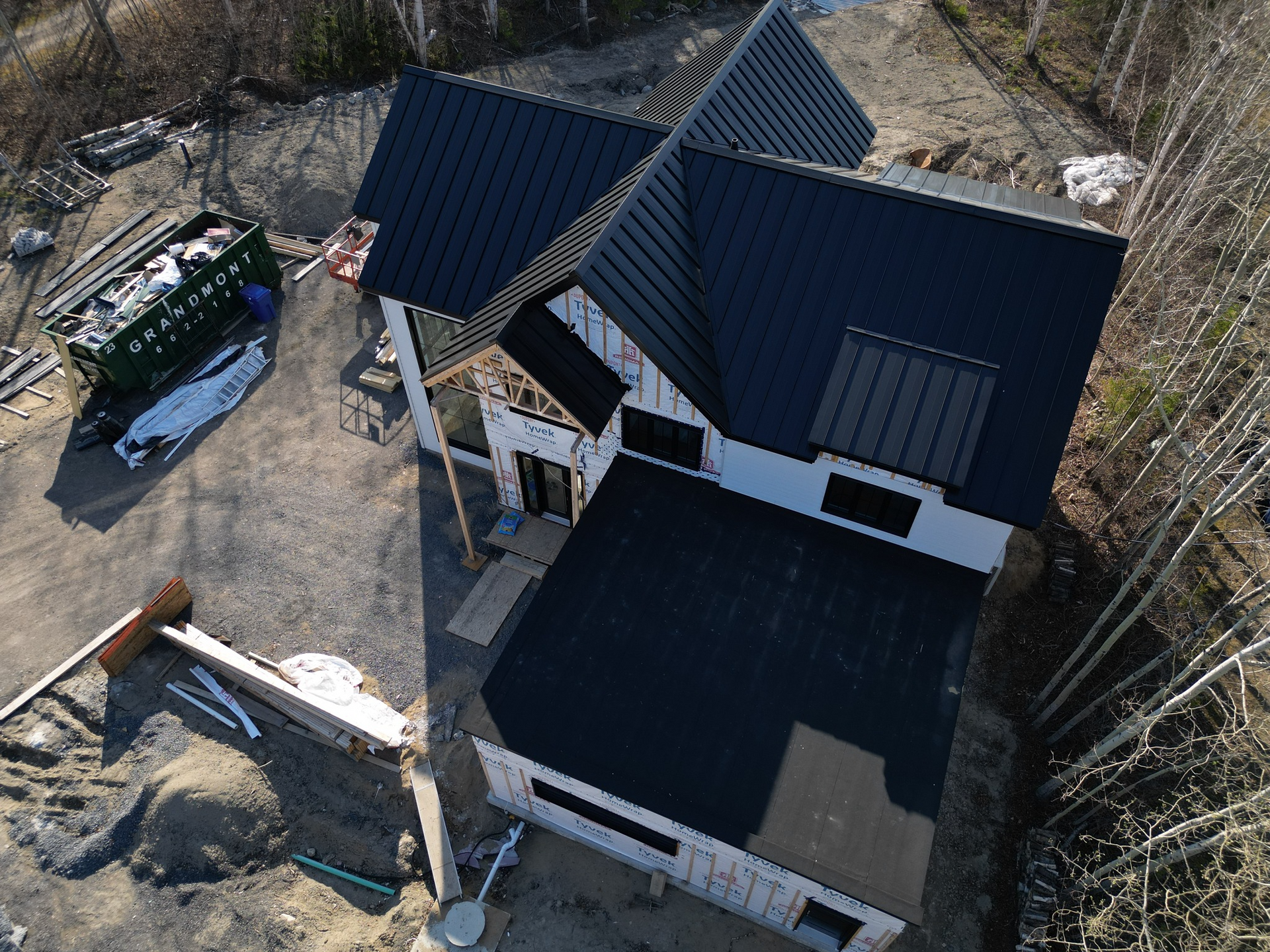 Vue aérienne d'une maison en construction, avec un toit sombre et un bardage blanc ; des débris de construction sont visibles.