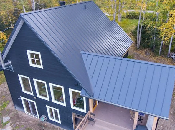 Maison gris foncé avec toit en métal assorti, encadrements de fenêtres blancs et porche, dans un cadre boisé.