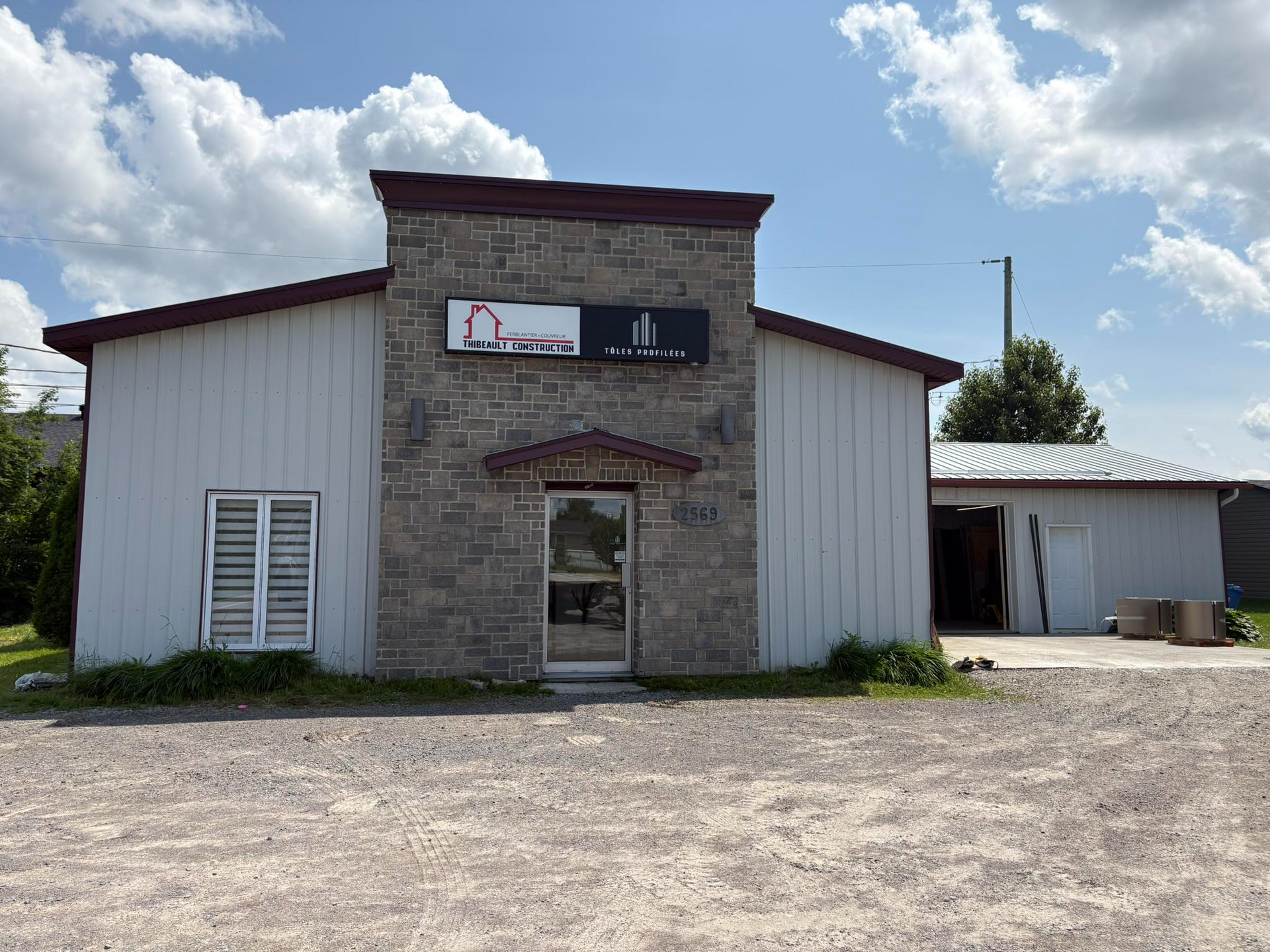 Extérieur d'un bâtiment de plain-pied avec une façade en pierre, un bardage gris et un parking en gravier sous un ciel bleu.