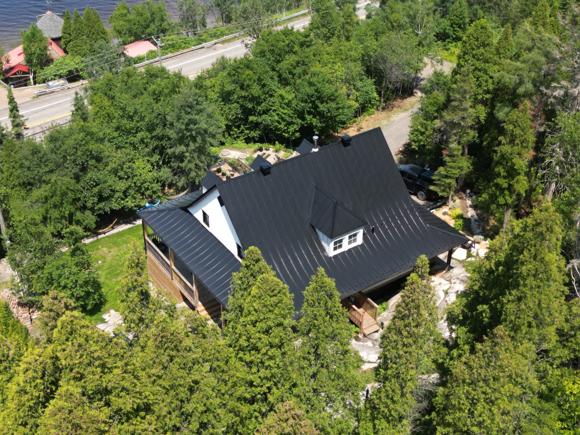 Vue aérienne d'une maison au toit en métal noir, entourée d'arbres, près d'une route et d'un point d'eau.