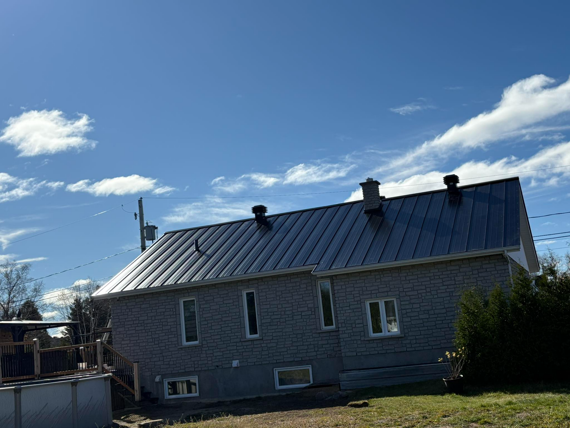 Maison grise au toit en métal sombre se détachant sur un ciel bleu, cheminée, fenêtres et petite cour.