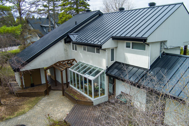 Maison à deux étages avec un toit en métal noir, une véranda vitrée et une terrasse en bois, entourée d'arbres.