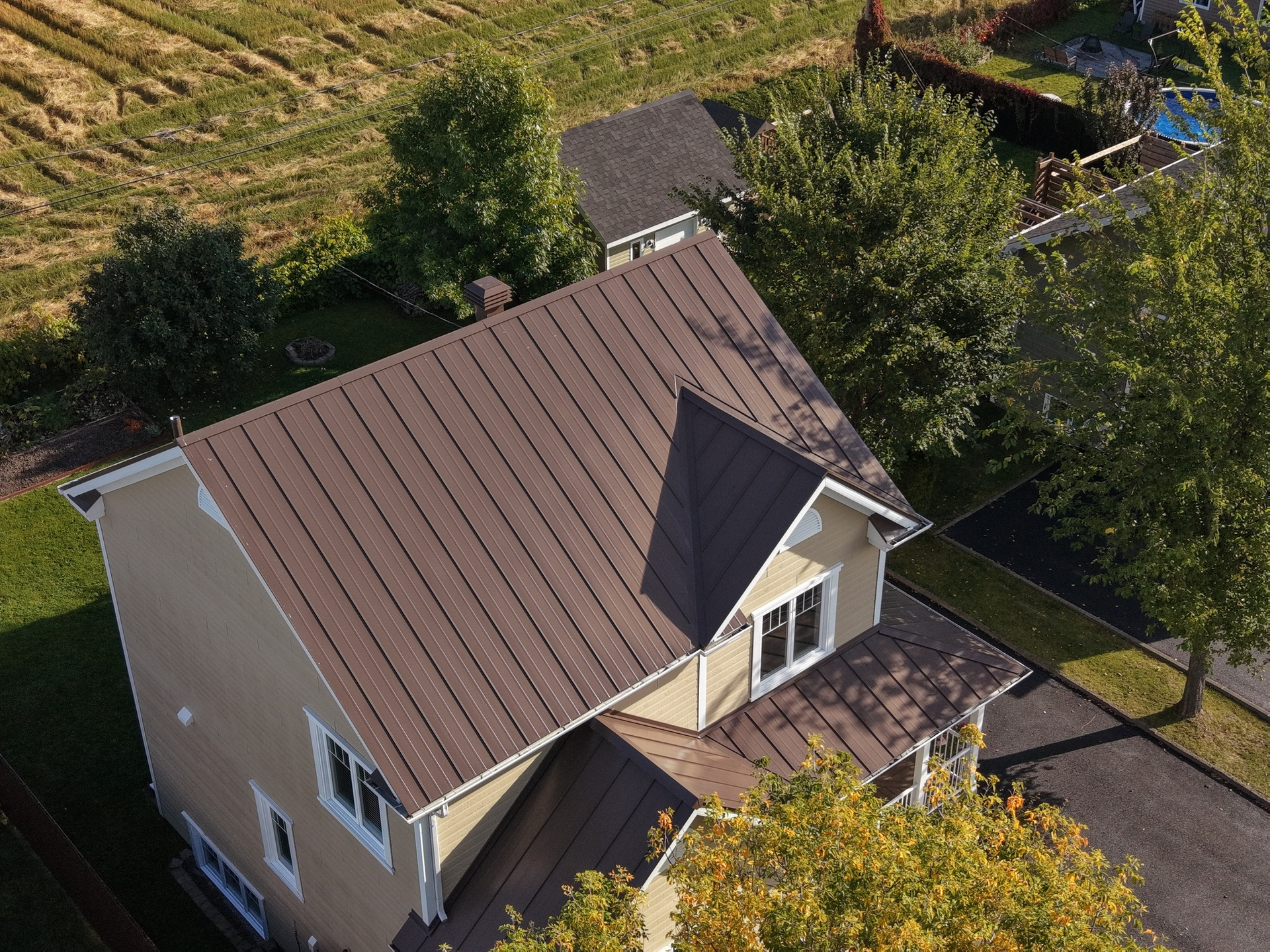 Vue aérienne d'une maison beige à deux étages avec un toit en métal marron, entourée d'arbres et d'une allée.