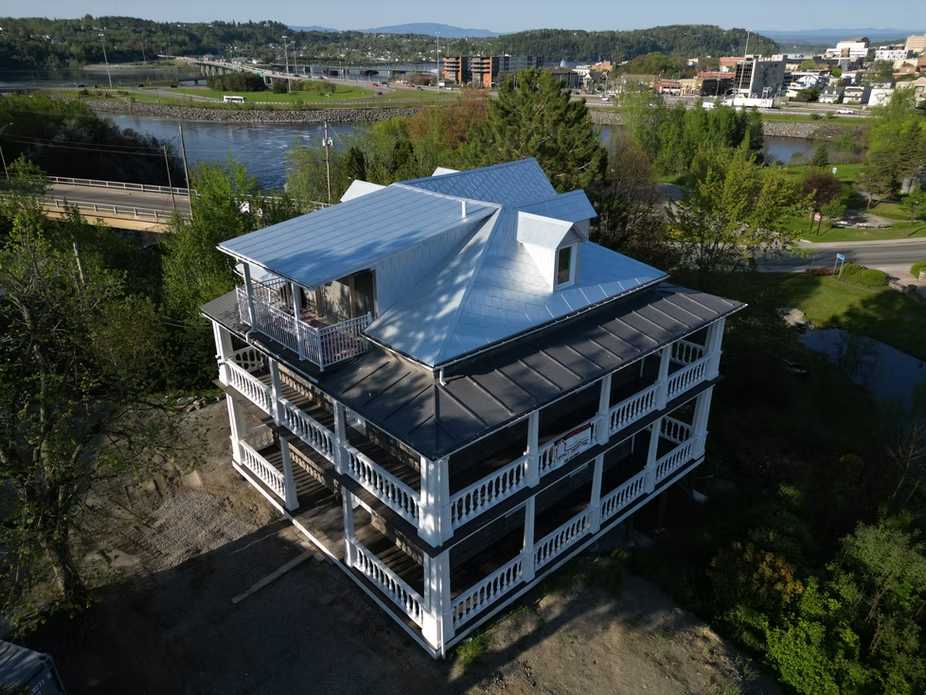 Maison blanche de trois étages avec balcons faisant le tour de la façade, toit en métal. Ville et rivière en arrière-plan.