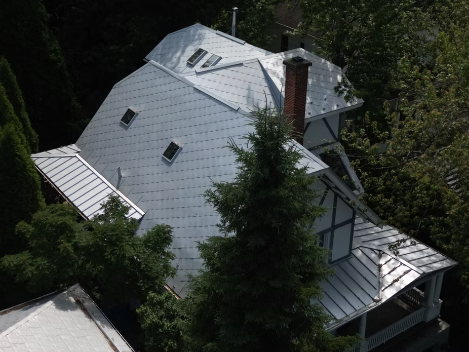 Maison avec un toit en métal brillant, des lucarnes et une cheminée en briques, entourée d'arbres.