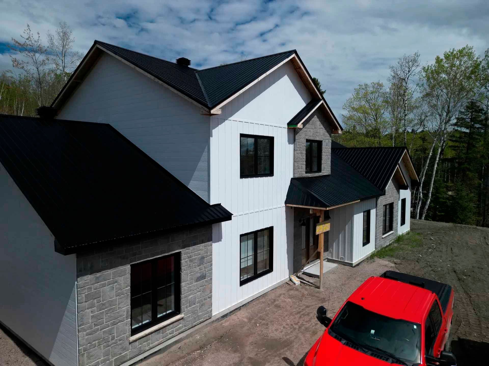 Maison à deux étages avec toit noir, bardage blanc, éléments en pierre et camion rouge dans l'allée.