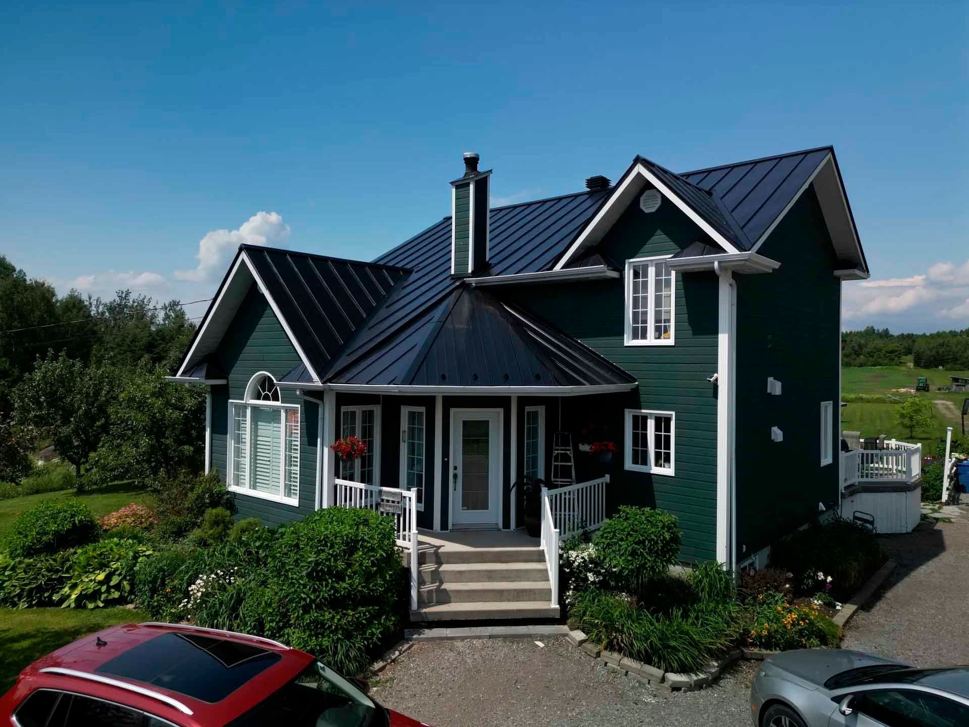 Maison verte au toit noir et aux boiseries blanches sous un ciel bleu, cour avant avec voitures garées.