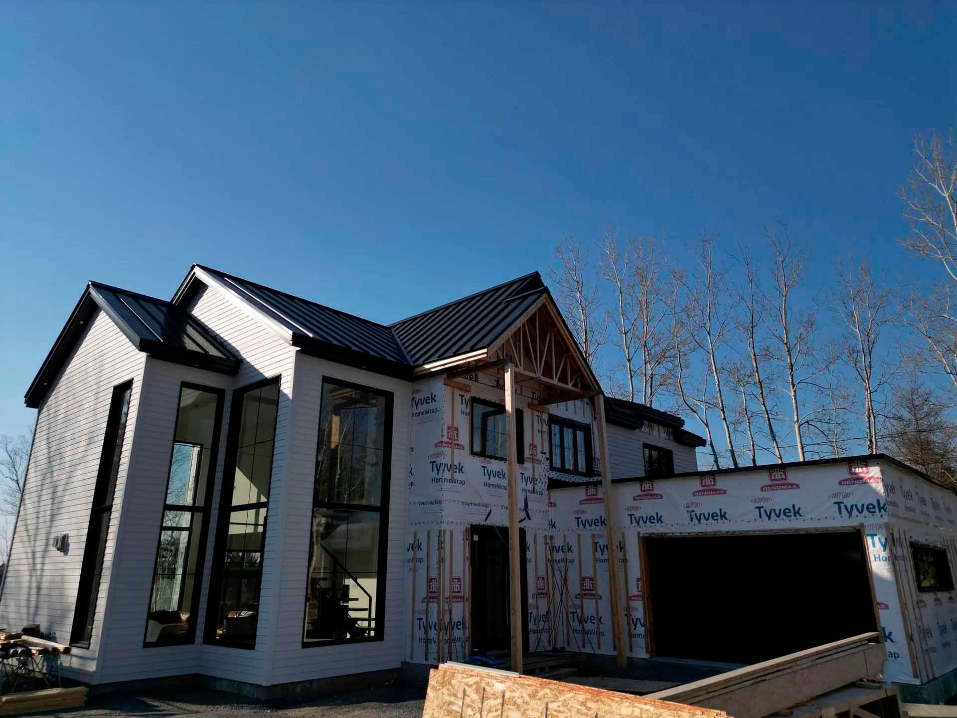 Maison à deux étages en construction, avec toit noir, bardage blanc, grandes fenêtres et membrane Tyvek. Ciel bleu en arrière-plan.