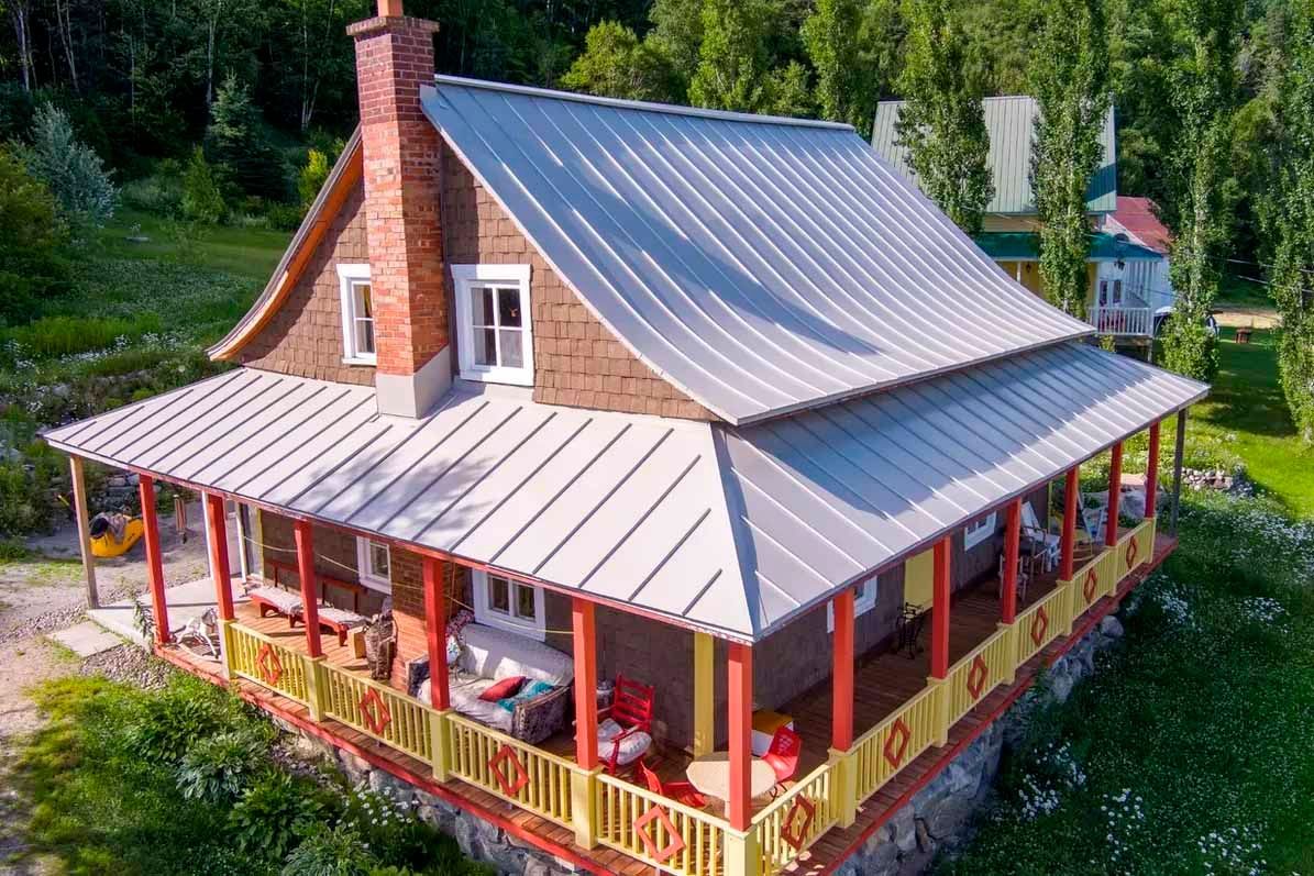 Maison rustique avec toit en métal, véranda enveloppante et cheminée en pierre.