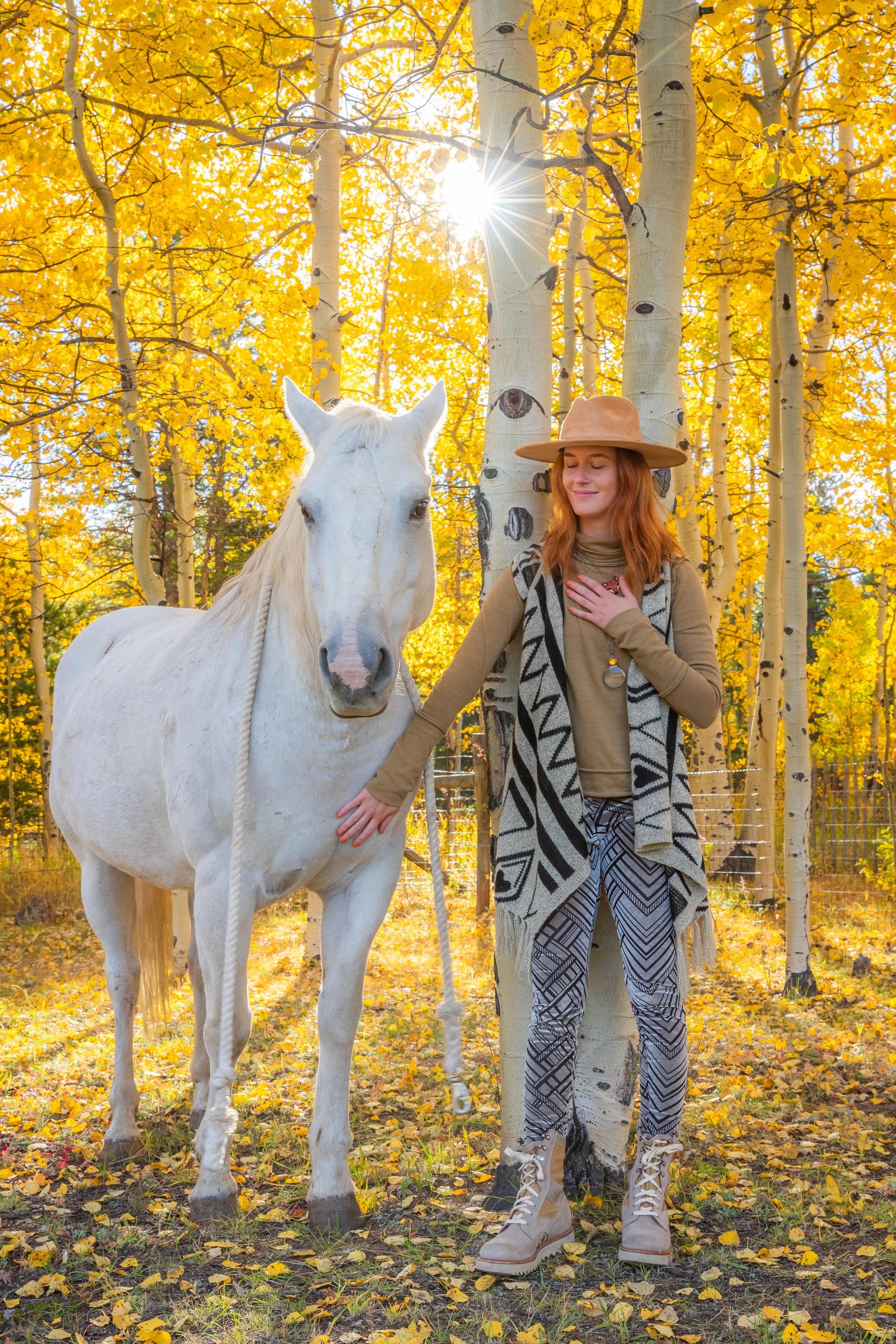 A person with reddish hair wearing a hat and patterned clothes sits on a wooden bench outdoors in a sunlit area.