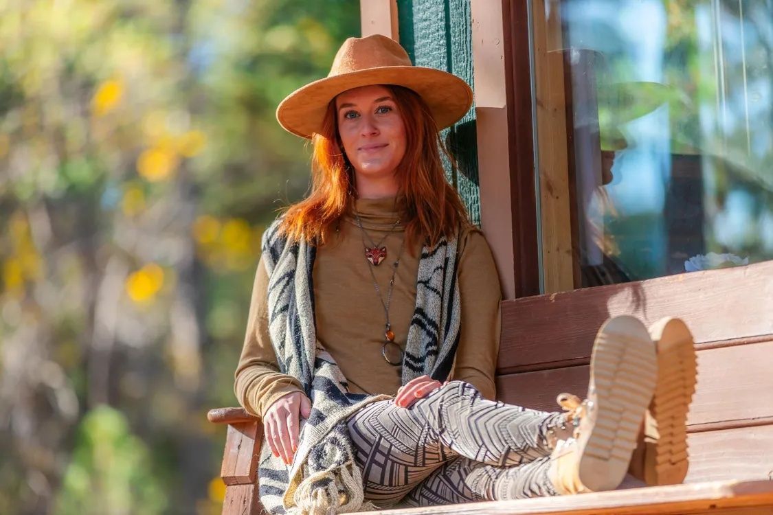 A person with reddish hair wearing a hat and patterned clothes sits on a wooden bench outdoors in a sunlit area.