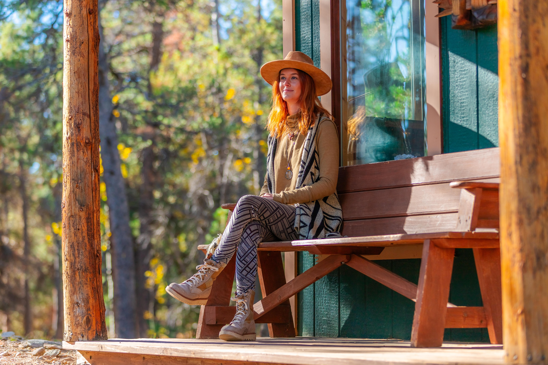 A person with reddish hair wearing a hat and patterned clothes sits on a wooden bench outdoors in a sunlit area.