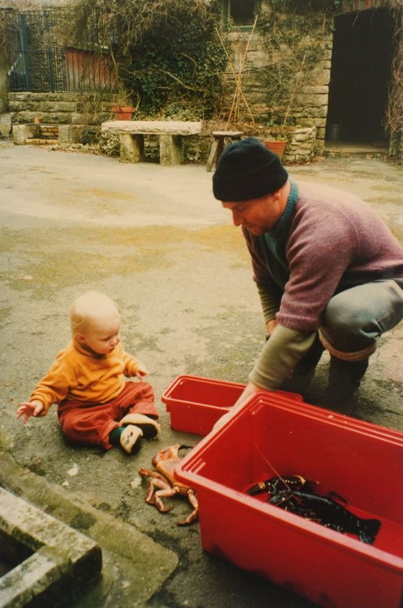 A baby watches a man near a red bin containing a lobster and octopus in an outdoor courtyard.
