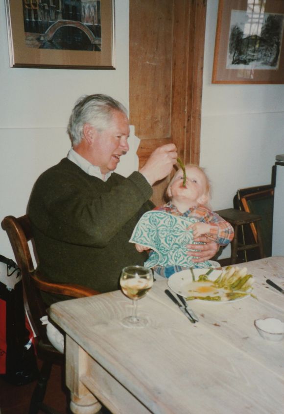Man feeding a child from a table. Man seated, holding child, offering green food. Table has asparagus, wine glass.